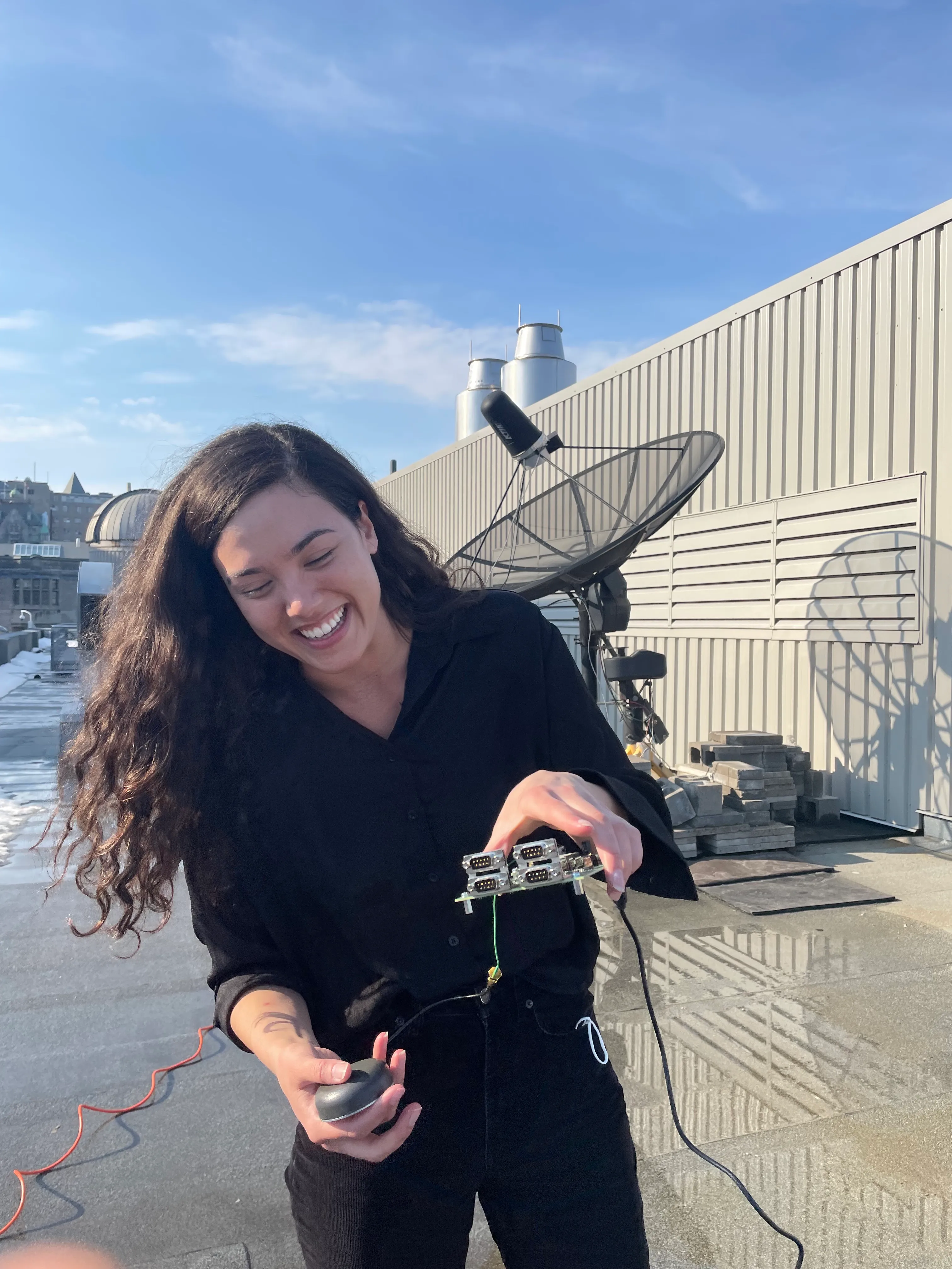 A researcher holding a small receiver on a sunny rooftop next to a satellite dish.