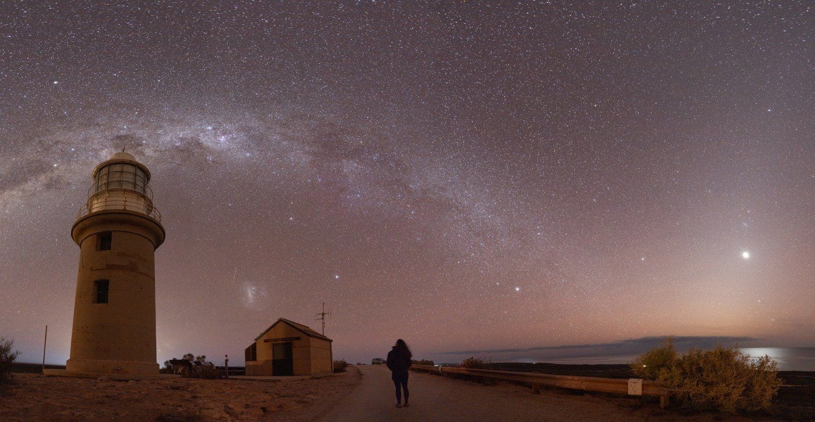 A lighthouse and a lone figure on a road beneath the arc of the Milky Way at night